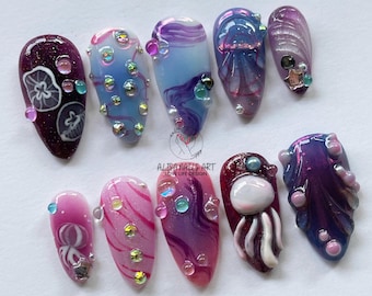 May include: Set of ten false nails with a sea-themed design. The nails are painted in shades of purple, blue, and pink with various sea creatures and designs, including jellyfish, seashells, and bubbles. Some nails are adorned with rhinestones.