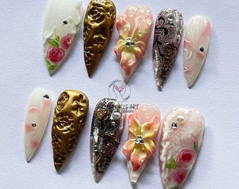 May include: A collection of ten long, pointed artificial nails. Designs include gold textured, floral, and abstract patterns. Some nails feature rhinestones and 3D floral embellishments. The color palette includes white, gold, pink, and black.