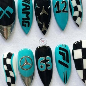 May include: A set of artificial nails with a racing theme. Designs include the AMG logo, the number 12, a Mercedes-Benz emblem, the number 63, and a checkered flag pattern. The nails are painted in black, teal, and silver.