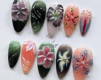 May include: A collection of ten artificial nails with various designs. Each nail features a different color scheme and design, including 3D floral embellishments, gradients, and abstract patterns. The nails are in an oval shape and are arranged on a light surface.