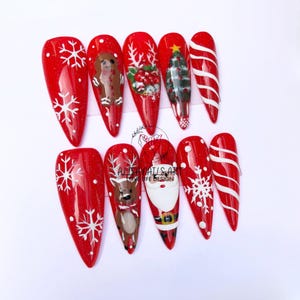 May include: Set of ten red stiletto false nails with white snowflake and Christmas-themed designs. The designs include a reindeer, Santa Claus, a Christmas tree, and candy canes.