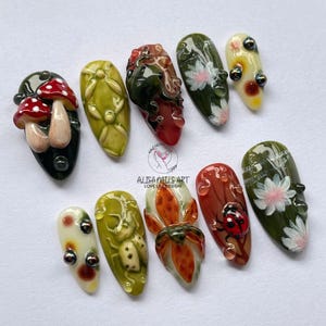 Könnte beinhalten: Zehn künstliche Fingernägel mit verschiedenen Designs, darunter Pilze, Blumen und Marienkäfer, mit glänzendem Finish. Die Farbpalette umfasst Rot, Grün, Weiß und Gelb. Der Text "ALISA NAILS ART LOVE LIFE DESIGN" ist ebenfalls sichtbar.