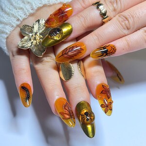 Könnte beinhalten: Eine Hand mit langen, mandelförmigen Nägeln, die aufwendige Nagelkunst zeigen. Designs umfassen Blätter, Blumen, ein Spinnennetz und Insekten, alles in Orange-, Braun- und Goldtönen. Die Hand trägt auch mehrere goldene Ringe.