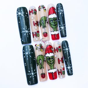 May include: Set of ten Grinch-themed Christmas nail art designs. The nails are long and have a green, white and red colour scheme. The designs feature the Grinch, a Christmas tree, and the words "Ho Ho Ho".