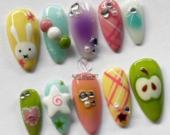 Puede incluir: Conjunto de diez uñas postizas de colores con varios diseños, incluyendo un conejo, una estrella, un patrón de cuadros, una manzana y un corazón. Las uñas están decoradas con diamantes de imitación y otros adornos.