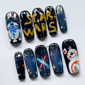 Puede incluir: Un conjunto de diez uñas artificiales con temática de Star Wars. Los diseños incluyen R2-D2, BB-8, las palabras "STAR WARS" en amarillo, un diseño de galaxia y sables de luz. Las uñas están pintadas en negro, azul, blanco y amarillo.