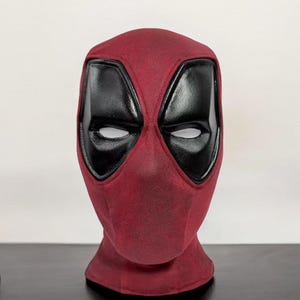 May include: A burgundy-colored Deadpool mask with black eye coverings and white eye openings. The mask is made of a textured fabric and is designed to cover the entire head. The mask is a cosplay item.