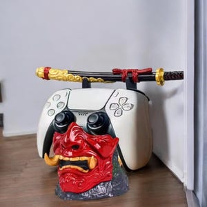 May include: A white gaming controller stand featuring a red and black Oni mask base. A decorative katana rests on top. The controller has black joysticks and button details. The katana has gold and red accents.
