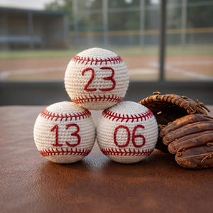 Puede incluir: Tres pelotas de béisbol de ganchillo color crema con detalles de costura rojos. La pelota superior tiene el número 23, mientras que las dos inferiores muestran los números 13 y 06 bordados en rojo. Un guante de béisbol de cuero marrón.