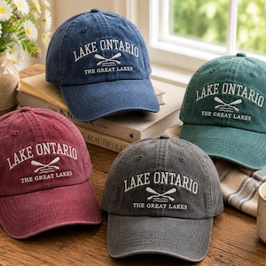 May include: Four baseball caps in blue, green, maroon, and charcoal gray. Each cap features white embroidered text reading LAKE ONTARIO and THE GREAT LAKES, with a graphic of two crossed oars above wavy lines centered on the front panel.