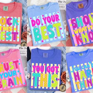 Pode incluir: Uma coleção de seis camisetas coloridas com frases motivacionais. Cada camiseta apresenta uma mensagem diferente em uma fonte vibrante e lúdica, incluindo "ROCK THIS TEST" e "YOU CAN DO HARD THINGS". As camisetas estão em várias cores, como rosa, azul e roxo.