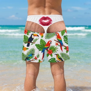 May include: White swim shorts featuring a tropical pattern of colourful parrots, toucans, and hibiscus flowers. The waistband displays a prominent red lip print centred at the back. The design includes various green palm leaves and foliage.