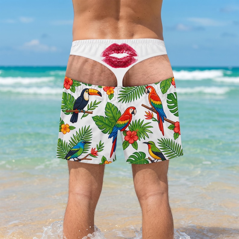 May include: White swim shorts featuring a tropical pattern of colourful parrots, toucans, and hibiscus flowers. The waistband displays a prominent red lip print centred at the back. The design includes various green palm leaves and foliage.