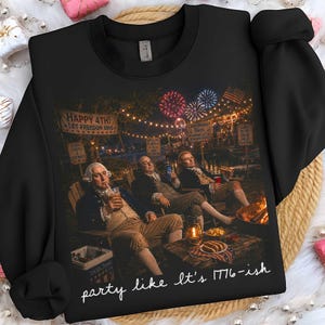 Może przedstawiać: Czarna bluza z nadrukiem przedstawiającym historyczne postacie świętujące 4 lipca z fajerwerkami i ogniskiem. Tekst na bluzie brzmi "HAPPY 4TH! LET FREEDOM RING" i "party like it's 1776-ish."