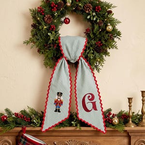 May include: A decorative fabric hanging featuring a light green and white pinstripe pattern with a red scalloped edge. One side displays an embroidered nutcracker figure, while the other side features a large, embroidered red letter G in script.