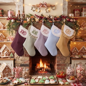 May include: Five personalised Christmas stockings in burgundy, sage green, lavender, light blue, and mustard yellow, each featuring a white cuff with a ruffled edge and embroidered initials, hanging in a row above a lit fireplace with holiday decor.