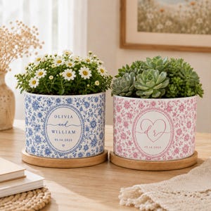 May include: Two cylindrical ceramic planters on wooden saucers. The left planter features a blue floral pattern with the text OLIVIA and WILLIAM and 05.14.2025. The right planter has a pink floral pattern with a heart, the letters dr, and 05.14.2025.