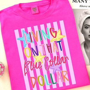 May include: A bright pink t-shirt with the text "LIVING ON THAT Blue Collar DOLLAR" in colorful, playful lettering. The design includes stars and vertical stripes. A small pink tag with text is visible at the top.