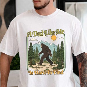 Può includere: T-shirt bianca con una grafica di Bigfoot che cammina in una scena di foresta, con montagne e un sole. Il testo "A Dad Like Me Is Hard To Find" è in verde e giallo.