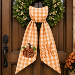 May include: A decorative fabric door hanger featuring an orange and white gingham pattern. The hanger has two long tails, one embroidered with a pumpkin featuring a leopard print pattern and green leaves, and the other with a large orange letter M.