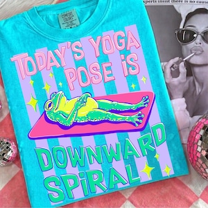 Może przedstawiać: Turkusowa koszulka Comfort Colors z napisem "TODAY'S YOGA POSE IS DOWNWARD SPIRAL" w kolorze różowym i zielonym. Grafika przedstawia kreskówkową żabę w pozycji jogi na różowej macie z żółtymi gwiazdami.