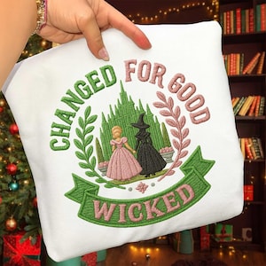 May include: White sweatshirt with an embroidered design featuring two figures in front of a green castle. The text reads "CHANGED FOR GOOD" and "WICKED" in green and pink. The design is surrounded by green leaves and pink branches.