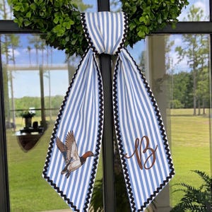 May include: A decorative fabric door hanger featuring light blue and white vertical stripes with a black zigzag trim. One side displays a flying mallard duck illustration, while the other side features a large, brown, cursive letter B monogram.