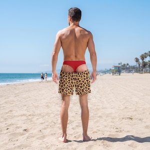 May include: Swim shorts featuring a bold leopard print pattern in shades of brown, black, and tan. The shorts have a standard elastic waistband and a relaxed fit, worn over a solid red thong-style undergarment, providing a distinct contrast.