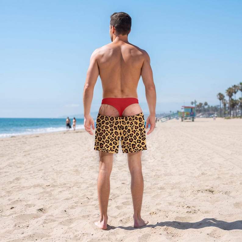 May include: Swim shorts featuring a bold leopard print pattern in shades of brown, black, and tan. The shorts have a standard elastic waistband and a relaxed fit, worn over a solid red thong-style undergarment, providing a distinct contrast.