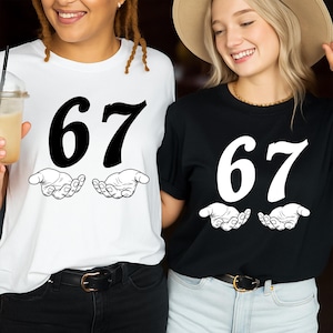 Puede incluir: Dos camisetas, una blanca y otra negra, cada una con el número "67" en negrita en blanco y negro, respectivamente. Debajo del número, un gráfico de dos manos abiertas está impreso. Las camisetas son usadas por dos personas.
