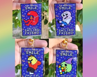 May include: Four Smiling Friends keychains. Each keychain has a different character on a blue background with the words "Alan makes me SMILE", "Pim makes me SMILE", "Charlie makes me a SMILE", and "Glep makes me SMILE". Each keychain has a gold-colored clasp.