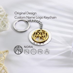 Pode incluir: Um chaveiro de ouro e prata com um logotipo de nome personalizado. O logotipo é um design geométrico com o nome "NORA" escrito dentro. O chaveiro está sobre um fundo branco.