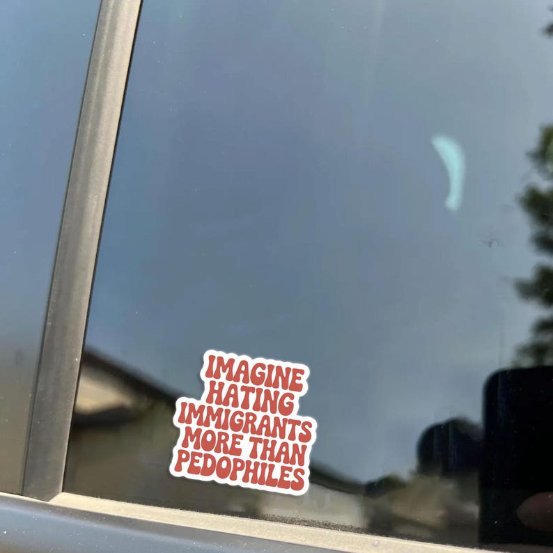 May include: A red and white sticker with the text "IMAGINE HATING IMMIGRANTS MORE THAN PEDOPHILES" is affixed to a car window. The sticker is rectangular with rounded corners and is positioned on the lower right side of the window.