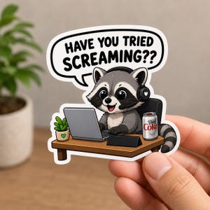 May include: A die-cut sticker featuring a cartoon raccoon wearing a headset, sitting at a desk with a laptop, a small potted plant, and a can of Diet Coke. A speech bubble above the raccoon contains the black text, HAVE YOU TRIED SCREAMING??