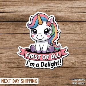 May include: A white unicorn sticker with a rainbow mane, a golden horn, and a pink flower. The sticker has the text "FIRST OF ALL I'm a Delight!" on a pink banner. The sticker is on a wooden background.