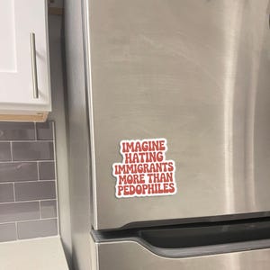 May include: A red and white sticker with the text "IMAGINE HATING IMMIGRANTS MORE THAN PEDOPHILES" is affixed to a stainless steel refrigerator door. The sticker is rectangular with rounded corners.