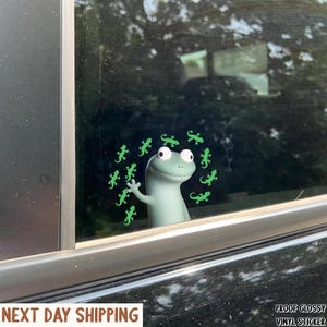 May include: A green vinyl sticker featuring a cartoon gecko waving from a car window, encircled by smaller green gecko illustrations. The sticker has a glossy finish. The text "PROOF GLOSSY VINYL STICKER" is visible.