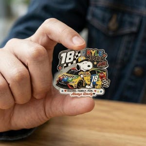 May include: A colorful pin featuring Snoopy in a racing suit next to a race car with the number 18. The pin has the text "KYLE BUSCH" and "Always Rowdy". The pin is held in a person's hand.