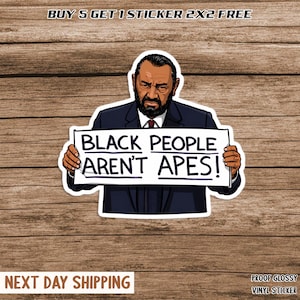 May include: A glossy vinyl sticker featuring a cartoon illustration of a man holding a sign. The sign reads "BLACK PEOPLE AREN'T APES!". The sticker is white with a black outline and is set against a wood grain background.