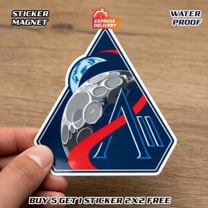 Könnte beinhalten: Ein dreieckiger Aufklebermagnet mit Weltraum-Design. Er hat einen blauen Hintergrund, einen stilisierten Mond und den Buchstaben "A". Die Wörter "STICKER MAGNET" und "WATER PROOF" sind aufgedruckt.