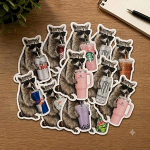 May include: A collection of die-cut stickers featuring raccoons wearing black sunglasses, each holding a different beverage including a Diet Coke can, a pink Starbucks cup, a Monster energy drink, a Red Bull can, and pink Stanley tumblers.