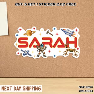 May include: A white sticker with the name "SARAH" in red, surrounded by space-themed illustrations. The sticker includes a planet, rocket, astronaut, and other space-related graphics. The text "BUY 5 GET 1 STICKER 2X2 FREE" is at the top.