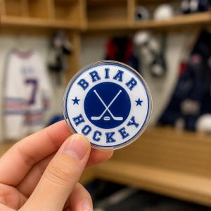 May include: A round, clear acrylic hockey puck with the words "BRIAR HOCKEY" in blue and white. The center features crossed hockey sticks and a puck. Two blue stars are on either side of the text.
