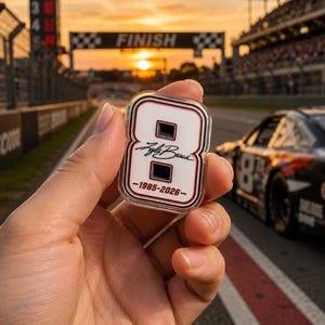 May include: A clear acrylic pin shaped like the number eight, with a white background and black and red trim. The pin features the name "Kyle Busch" and the years "1985-2026". A race car is visible in the background.
