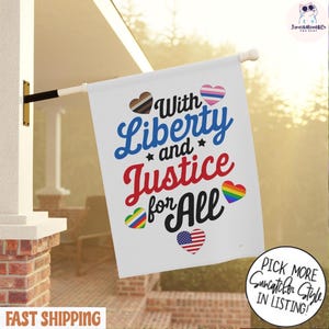 Könnte beinhalten: Eine weiße Flagge mit dem Schriftzug "With Liberty and Justice for All" in Blau und Rot. Die Flagge zeigt herzförmige Grafiken in verschiedenen Farben und Mustern, darunter ein Regenbogenherz und ein Herz in den Farben der US-Flagge.