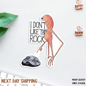 May include: A glossy vinyl sticker featuring a whimsical illustration of a creature with the text "I DON'T LIKE THIS ROCK." The creature is pointing at a grayscale rock. The sticker has a white border.