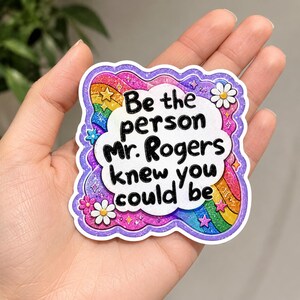 Peut inclure: Un autocollant d&eacute;coup&eacute; avec un centre en forme de nuage blanc portant le texte noir 'Be the person Mr. Rogers knew you could be'. La bordure est un motif arc-en-ciel scintillant et vibrant avec de petites fleurs roses et blanches et des &eacute;toiles sur un fond violet.