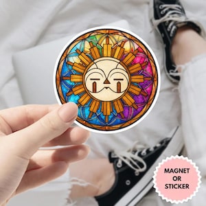 Puede incluir: Una pegatina o imán redondo con un diseño de sol de vidriera con una cara. El sol tiene un centro amarillo con rayos naranjas y un borde de arcoíris colorido. El texto "MAGNET OR STICKER" está en una pegatina rosa en la esquina inferior derecha.