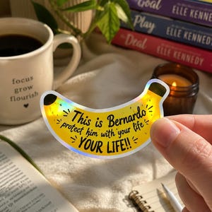 May include: A banana-shaped sticker with a holographic finish featuring the black text: This is Bernardo, protect him with your life, YOUR LIFE!! The sticker has a yellow base color with small star accents and a dark brown tip at the end.