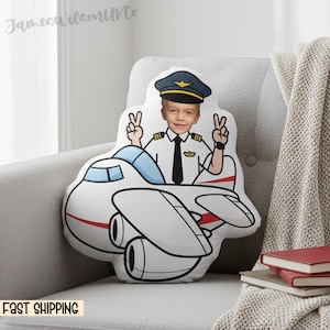 May include: A white airplane-shaped pillow with a cartoon pilot design. The pillow features a child's face in a pilot's uniform, giving peace signs. The pillow is on a gray chair with a blanket and books.